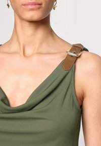 Olivgrön ärmlös topp med draperad halsringning, med en brun läderrem och detaljer i guld på axeln. Mjuk tygtextur.