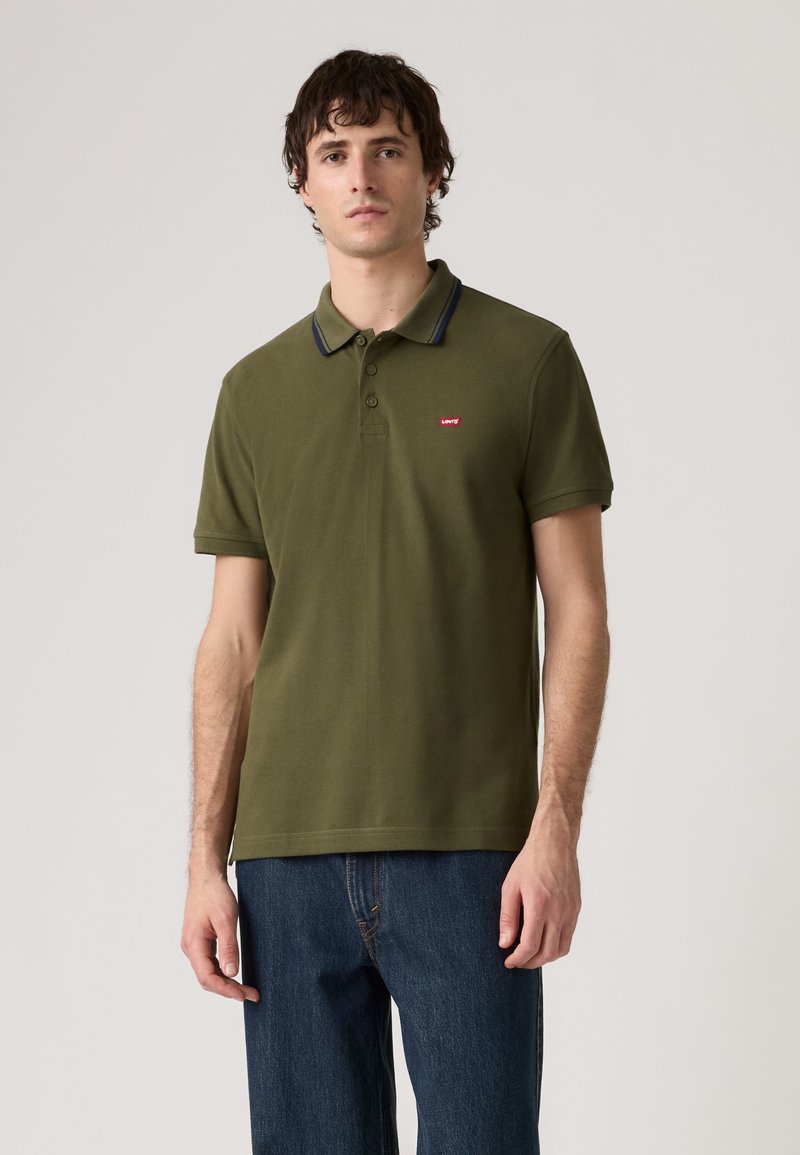 Homme portant un polo vert olive à manches courtes avec un bord foncé sur le col et un jean bleu, debout devant un fond clair uni.