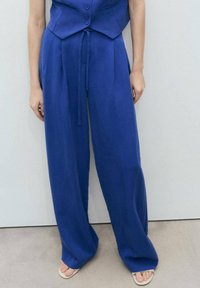 Leveled pants i en livlig blå farve med en glat tekstur. Har en bundet talje og en afslappet pasform, der supplerer en tætsiddende vest.