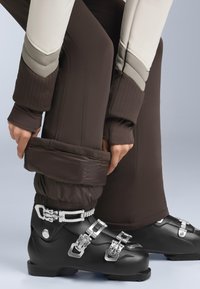 Fekete síbakancs ezüst csatokkal, barna, texturált sínadrággal párosítva, bordázott szegéllyel és bézs és szürke paneles kialakítással.