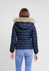 Námořnická péřová bunda s kapucí s umělým kožichem, s vodorovným prošíváním, zapínáním na zip vpředu a žebrovanými manžetami. Nošena přes světle modré džíny.