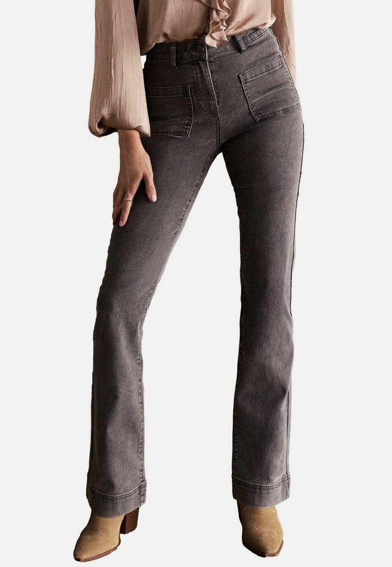 Jeans grii evazați din denim, cu buzunare frontale și dorsale, talie înaltă și margine brută. Purtați cu o bluză de culoare deschisă și cizme cu toc bej.