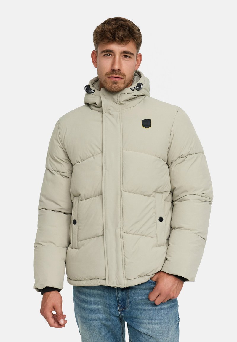 Beżowa kurtka puchowa z kapturem, z logo na piersi, pikowanymi sekcjami, napami oraz elastycznymi mankietami. Noszona na niebieskie dżinsy.