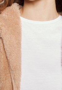 Femme portant un manteau beige en peluche par-dessus un t-shirt blanc à col rond, mettant en avant le cou et le haut de la poitrine.