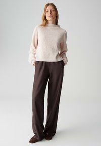 Maglione rosa chiaro con texture e maniche lunghe abbinato a pantaloni a gamba larga marrone scuro. L'outfit presenta una vestibilità morbida e un tessuto liscio.