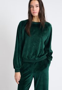 Felpa in velluto verde con vestibilità ampia, polsini e orlo a coste, e spalle scese, che presenta una texture morbida e liscia.