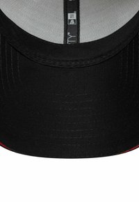 Gorra de béisbol negra con una corona estructurada y visera plana, que presenta una tela texturizada y un detalle de acento rojo a lo largo del borde.