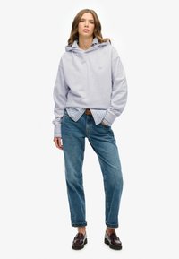 Sudadera lila con un ajuste relajado, superpuesta sobre una camisa de rayas azules y blancas. Combinada con jeans de mezclilla azules y mocasines marrones.
