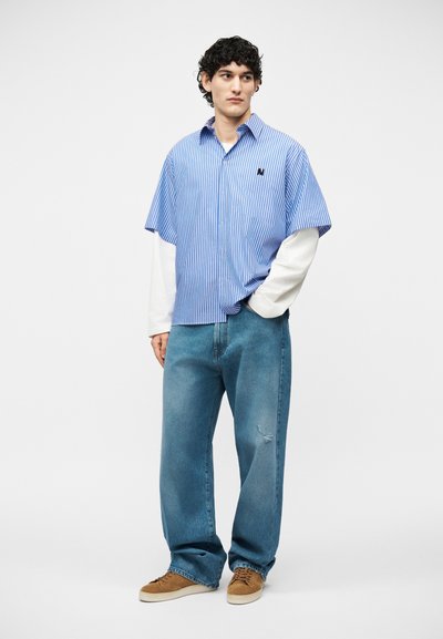 Jeune homme portant une chemise à manches courtes rayée bleue superposée à un t-shirt blanc à manches longues, un jean bleu ample déchiré et des baskets marron, debout les mains dans les poches.