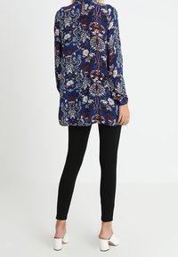 Marinblå tunika med vita blommiga och bladmönster, långa ärmar och rund halsringning; matchad med svarta leggings och vita klackar.