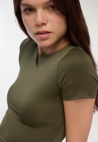 Oliwkowy top z krótkim rękawem, z okrągłym dekoltem. Gładki, dopasowany materiał oraz bezszwowy design podkreślają kształt. Subtelne, lśniące wykończenie.