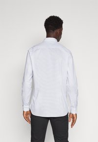Camicia bianca a maniche lunghe con un sottile motivo geometrico, collo con bottoni e polsini con un bottone, abbinata a pantaloni neri.