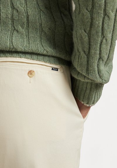 Πράσινο πουλόβερ με πλέξη καλωδίων και ριμπ μανσέτες σε συνδυασμό με κρέμ παντελόνι που διαθέτει κλείσιμο με κουμπί και μικρή ετικέτα μάρκας.