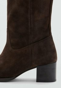 Bota de ante marrón hasta la rodilla con forma cónica, que presenta un robusto tacón bloque y detalles de costura sutiles. Textura suave.