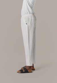 Persona vistiendo pantalones blancos holgados y sandalias negras sin talón, de pie contra un fondo neutral liso.