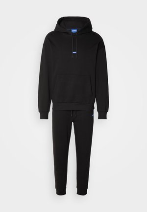 Hoodie noir avec une poche avant et une capuche à cordon, associé à un pantalon de jogging ajusté assorti. Présente un détail de logo bleu.