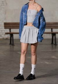 Femme portant un crop top gris et une jupe plissée, veste en jean bleu décolletée, chaussettes blanches avec chaussures noires, debout à l'intérieur.