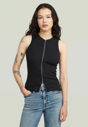 Frau mit langen dunklen Haaren, trägt ein ärmelloses schwarzes Top mit Reißverschluss vorne und blaue Jeans, steht mit einer Hand in der Tasche vor einfarbigem Hintergrund.