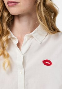 Camisa blanca de manga larga confeccionada con tejido ligero, que presenta un estampado de labios bordado en rojo en el lado izquierdo del pecho.
