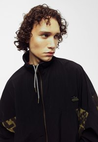 Chaqueta negra con un alto cuello, cierre delantero y tela con patrón de camuflaje en las mangas. Presenta un parche con un logo y detalle de cordón.