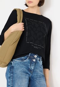 Zwart langarmshirt met grafische tekst, gecombineerd met blauwe spijkerbroek met een sluiting met twee knopen. De drager houdt een beige, gewatteerde tas vast.