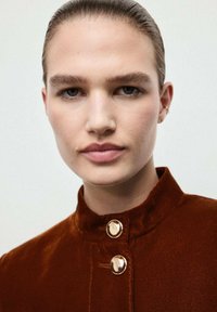 Giacca in velluto marrone con colletto alto, caratterizzata da due bottoni dorati sulla gola e una texture liscia. Toni della pelle morbidi e sfondo neutro.