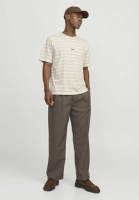 Camiseta a rayas beige y blanca con cuello redondo y logo, combinada con pantalones de tela marrones con pliegues y zapatos marrones de cordones. Lleva una gorra marrón.