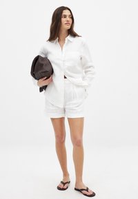 Hvid linned skjorte med knapper og en enkelt brystlomme, parret med hvide linned shorts. Model holder en struktureret brun taske.