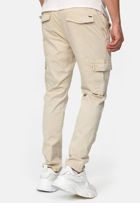 Beige cargo-housut, joissa on kapeampi istuvuus, useita taskuja ja hienovaraisia ompeluyksityiskohtia. Yhdistetty valkoisiin urheilukenkiin.