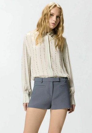 Giovane donna con lunghi capelli biondi che indossa una camicetta crema con borchie verticali e pantaloncini grigi a vita alta su uno sfondo semplice.