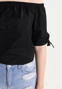 Top nero a spalle scoperte con maniche a palloncino corte, dotato di bottoni sul davanti e scollatura elastica. Abbinato a jeans blu strappati.