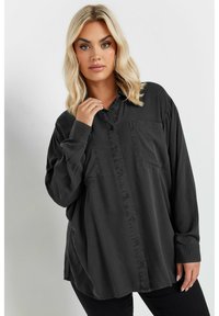 Camisa de botones negra de gran tamaño, confeccionada en un material suave, con un único bolsillo en el pecho y mangas largas con puños abotonados.