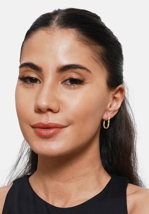 Gouden hoopoorbellen met een gestructureerd oppervlak, uitgerust met een scharniervergrendeling. Het model heeft donker haar, draagt een zwarte top en heeft een neutrale lipkleur.