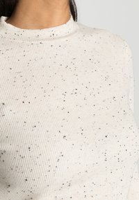 Top de malha canelada creme com discretas manchas pretas, gola alta e design justo. Tecido texturizado com riscas verticais.