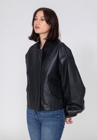 Schwarze Leder-Bomberjacke mit einem Reißverschluss vorne, übergroßen Ärmeln und einem dezenten Logo auf der Brust. Verfügt über Seitentaschen und eine glatte Textur.