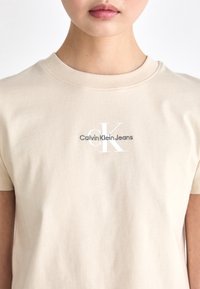 Persona che indossa una t-shirt beige di Calvin Klein Jeans con un logo bianco "CK" e testo nero sul petto, mostrata dalla nuca fino a metà torace.