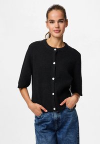 Svart strikket cardigan med rund hals, korte ermer og hvite knapper. Brukes over blå denimjeans og har et teksturert stoff.