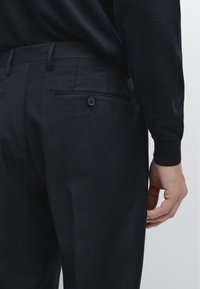 Dopasowane czarne spodnie o gładkiej fakturze, z tylną kieszenią zapinaną na guzik oraz szlufkami na pasek, widoczne od tyłu.