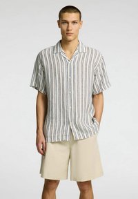 Camisa de manga corta, abotonada, con rayas verticales en tonos neutros. Hecha de un tejido liviano, de corte relajado. Combinada con pantalones cortos beige.