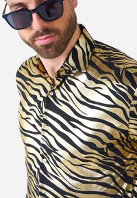 Chemise rayée or et noir avec une texture brillante, dotée d'un col classique et d'une fermeture à boutons. Des lunettes de soleil sombres complètent le look.
