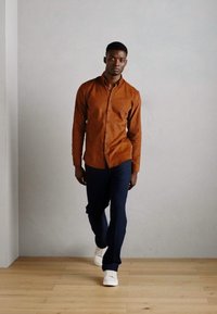 Cămașă cămilie, pantaloni bleumarin și adidași albi. Cămașa are o textură netedă, iar ținuta are o croială modernă.