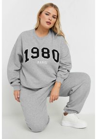 Sudadera gris con el texto negro "1980" y "PARÍS", con un corte relajado y puños acanalados. Combinada con pantalones de chándal grises a juego y zapatillas blancas.