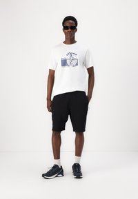 Hvit bomull T-skjorte med blå grafisk tekst og illustrasjon, kombinert med svarte shorts og mørke joggesko. Modell iført solbriller.