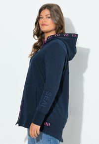 Donna che indossa una felpa con zip di colore blu navy, con simboli dell'infinito rosa sulla fodera del cappuccio e la scritta "SEAL" sulla manica, posta di profilo.
