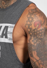 Grijze mouwloze shirt met een brede armsgat, voorzien van witte grafische tekst. Gedetailleerde tatoeages op de arm bevatten bloem- en schedelpuntens.
