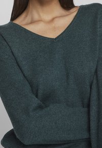 Pull en laine vert sarcelle foncé avec un col en V, présentant une texture douce et une coupe décontractée. Les manches sont légèrement surdimensionnées et resserrées aux poignets.