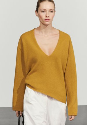 Vrouw draagt een mosterdgele trui met V-hals en witte broek, houdt een zwarte schoudertas vast tegen een effen lichte achtergrond.