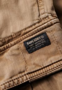 Tissu texturé marron avec une étiquette noire indiquant "SUPERDRY" cousue dans la couture, comportant du texte en blanc et en japonais. Perspective en gros plan.