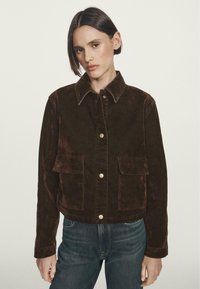 Kvinde med kort, mørkt hår iført en mørkebrun knapforret jakke med frontlommer og blå jeans, stående mod en ensfarvet lys baggrund.