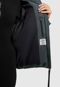 Hånd, der holder en åben mørkegrøn jakke, som viser den sorte foring, den lynlåslomme og mærket, der står "Et varmt kram lavet af stof."